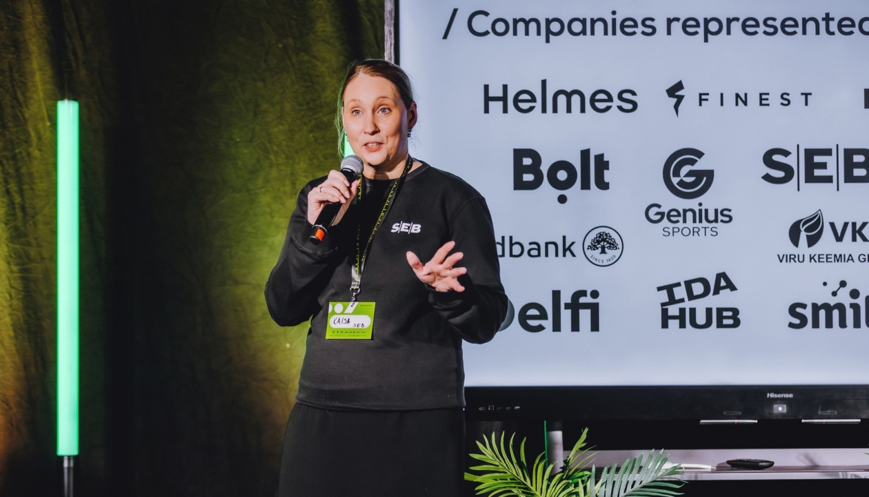 Woman presenting on a stage at kood/connect job fair in SEB hoodie with technology companies Bolt, Helmes, SEB, Delfi, Genius, VKG, Swedbank in background